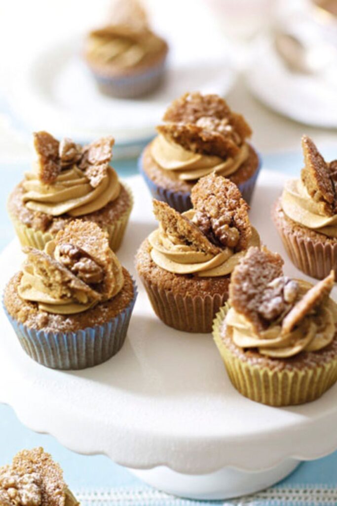 These coffee and walnut cupcakes are rich, nutty, and perfect for any coffee lover. They have a soft and fluffy texture, a delicious coffee flavour, and crunchy walnuts in every bite. Topped with smooth coffee buttercream and a classic butterfly decoration, they are great for afternoon tea, birthdays, or special occasions. In this recipe Mary Berry uses the all-in-one method, making it super easy and foolproof—just mix everything in one bowl and bake! Whether you’re making them for a gathering or just to enjoy with a cup of coffee, these cupcakes are a guaranteed hit. Ingredients Needed For the cupcakes: 2 tsp instant coffee granules 115g (4oz) butter (room temperature) or baking spread (at least 70% fat) 115g (4oz) caster sugar 115g (4oz) self-raising flour 1 tsp baking powder 2 large eggs 45g (1 1/2oz) walnut halves, finely chopped For the icing: 2 tsp instant coffee granules 350g (12oz) icing sugar, plus extra for dusting 175g (6oz) butter (room temperature) 6 walnut halves, halved lengthways How To Make Coffee and Walnut Cupcakes Prepare the oven and coffee: Preheat the oven to 180°C (fan 160°C/350°F/Gas 4). Line a 12-hole shallow bun tin with paper cases. In a small bowl, dissolve the coffee granules in 2 teaspoons of hot water, then set aside to cool. Make the cake batter: In a large bowl, combine the butter, caster sugar, self-raising flour, and baking powder. Add the eggs and the cooled coffee mixture. Using an electric hand whisk or a wooden spoon, beat until the mixture is smooth and creamy. Stir in the chopped walnuts. Bake the cupcakes: Divide the batter evenly between the paper cases. Bake for 15–20 minutes until the cupcakes are well-risen and firm to the touch. Transfer them to a wire rack and let them cool completely. Make the coffee icing: In a small bowl, dissolve the coffee granules in 2 teaspoons of hot water and let it cool. Sift the icing sugar into a large bowl. Add the butter and cooled coffee mixture, then beat until smooth and fluffy. Decorate the cupcakes: Using a sharp knife cut a small circle from the top of each cupcake and cut each piece in half to create “butterfly wings”. Spoon the icing into a piping bag fitted with a star nozzle and pipe a swirl into the centre of each cupcake. Place the reserved cake pieces on top to resemble wings, then add a walnut quarter between them. Lightly dust with icing sugar before serving. Recipe Tips Use soft butter: Let the butter sit at room temperature for 2–3 hours before baking to ensure a smooth mixture. Even baking: Fill the paper cases evenly so that all cupcakes rise at the same rate. Smooth icing: Sift the icing sugar before mixing for a lump-free, creamy texture. No piping bag? Simply spread the icing on top with a butter knife or spoon for an easy alternative. How To Store Leftovers Storing in the fridge: Place the cupcakes in an airtight container and store them in the fridge for up to 3 days. Bring them to room temperature before serving for the best texture. Freezing without icing: Store uniced cupcakes in a freezer-safe container for up to 3 months. Thaw them at room temperature before decorating. Freezing with icing: If freezing with icing, arrange the cupcakes in a single layer inside a container and freeze for up to 2 months. Allow them to thaw in the fridge overnight, then let them sit at room temperature before serving. Try More Recipes: Mary Berry Chocolate Cupcakes Mary Berry Ginger Cupcakes Mary Berry Lemon Cupcakes Mary Berry Carrot Cupcakes